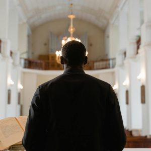 A priest stands before a congregation in a grand church, delivering a sermon with an open Bible.