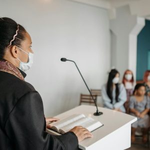 Female pastor delivering a sermon to a masked congregation indoors.